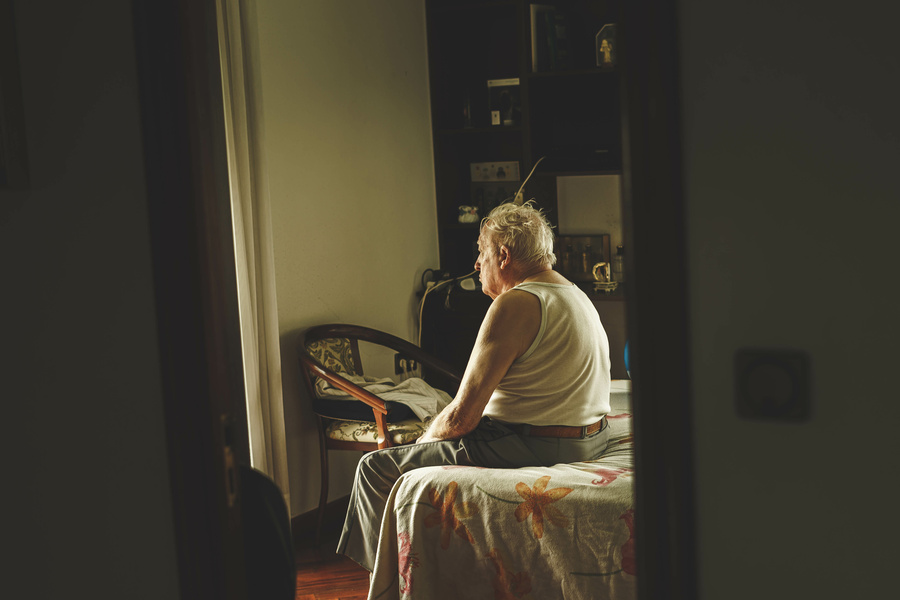 old man sitting at the foot of a bed seen from the setting of the room